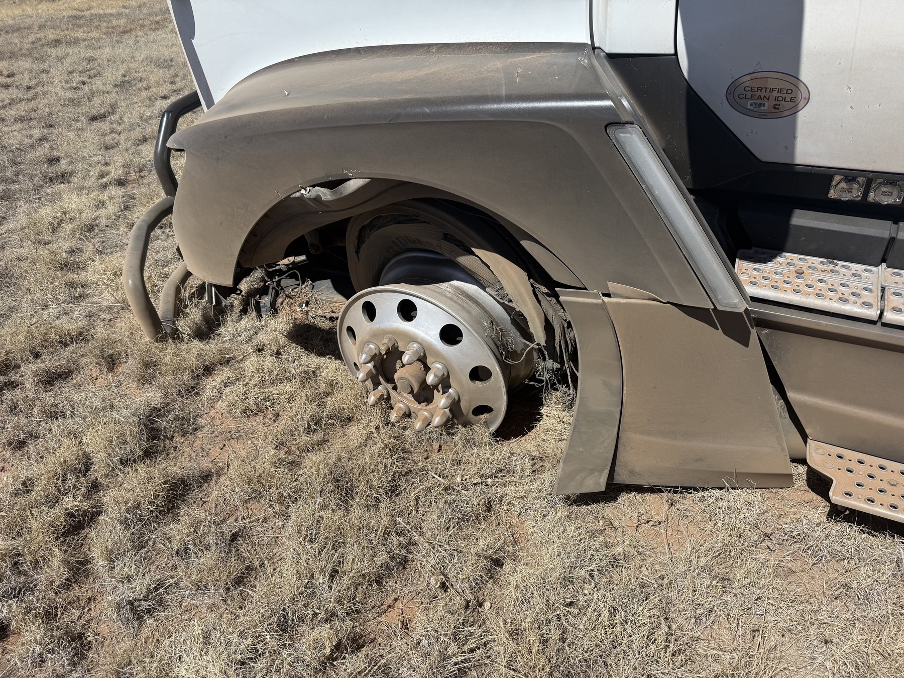 Truck E15767's steer tire destroyed to the bare rim after a blowout, the truck stopped in the grass beside the lane